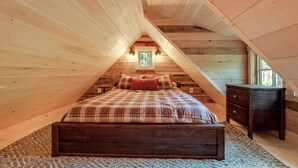 Un dormitorio acogedor en el ático con techo inclinado de madera, una cama con ropa de cama escocesa, una mesita de noche y un aparador.