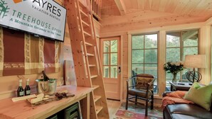 Interior acogedor de una cabaña con detalles de madera, una zona de comedor con una mesa dispuesta para dos personas, una silla y vistas al exterior a través de amplios ventanales.