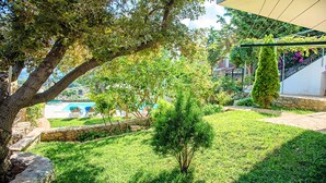 A lush garden with a stone pathway, a small tree, and a view of a pool and buildings in the background. - Grand View Villa | Heated Pool (Georgioupoli)