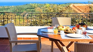 A dining table set outdoors with a view of the sea and hills. - Grand View Villa | Heated Pool (Georgioupoli)