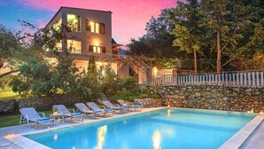 A swimming pool with lounge chairs, a stone wall, and a two-story building with balconies. - Grand View Villa | Heated Pool (Georgioupoli)