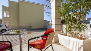 Uma área externa com assentos, uma mesa redonda e duas cadeiras, um vaso com plantas e um edifício ao fundo.