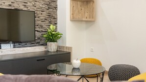 Un salón moderno con una televisión de pantalla plana montada en una pared con textura de piedra, un mueble de madera, una mesa de centro de vidrio con un jarrón blanco y una manzana, y un taburete amarillo de punto.