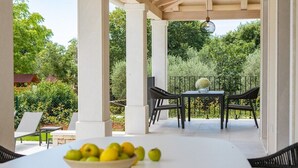 A covered patio area with a table and chairs, a bowl of fruit, and a view of lush greenery. - Modernly designed villa located near Pula (Manjadvorci)