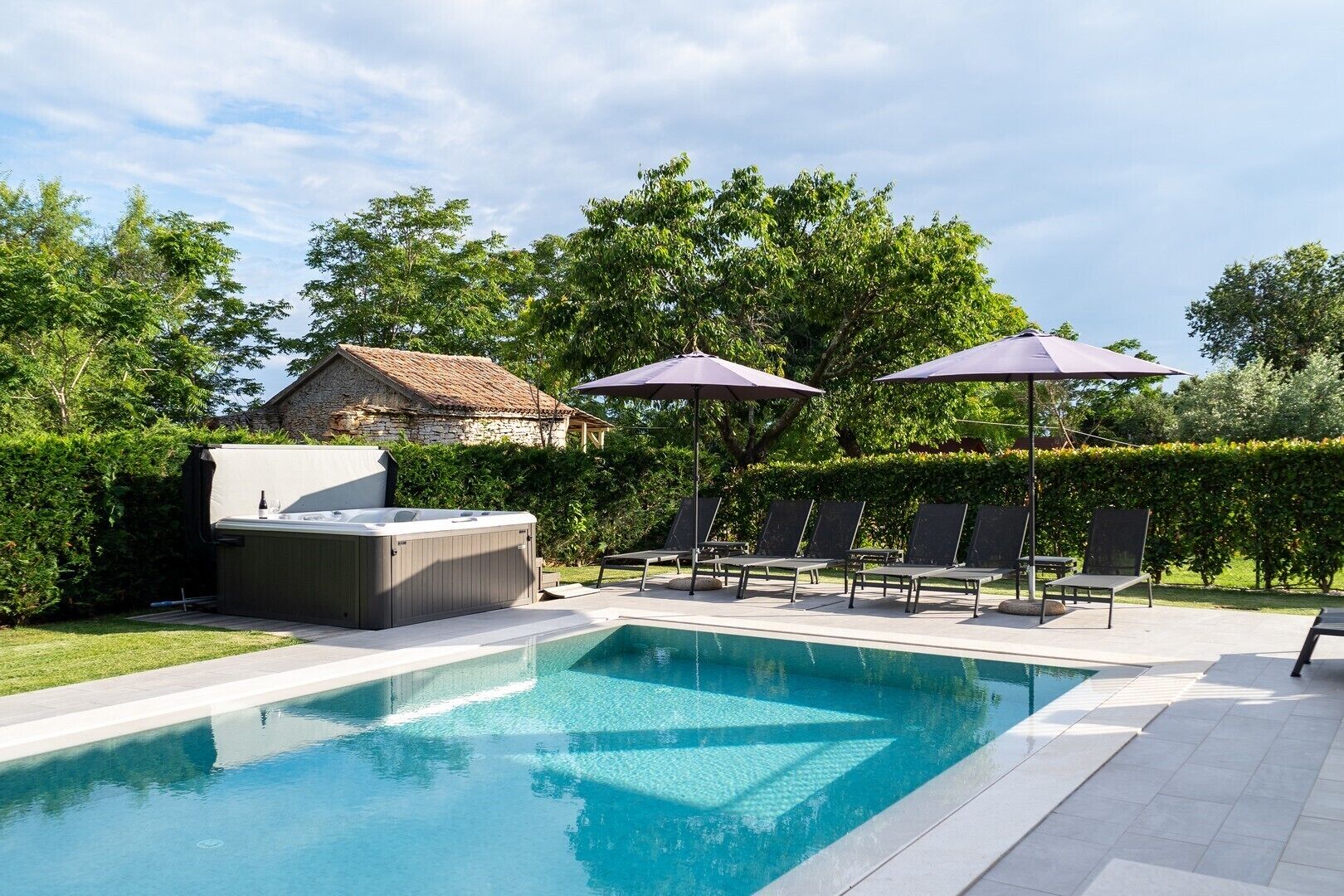 Piscina com espreguiçadeiras e guarda-sóis, em meio a vegetação e um edifício de pedra.