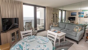 Un soggiorno con televisione, tavolo da pranzo con piatti, un divano e vista sul mare attraverso ampie finestre.