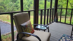 Un espace extérieur aménagé, abrité et doté de sièges confortables, avec une chaise rembourrée et une petite table, offrant une vue sur un paysage verdoyant et luxuriant.