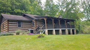 Une cabane en rondins dotée d’un porche et de fondations en pierre, entourée d’une cour verdoyante et d’arbres.