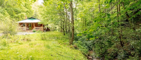 A cabin in a forested area with a porch and a small stream nearby.