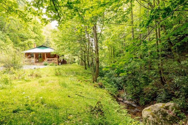A cabin in a forested area with a porch and a small stream nearby.