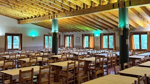 Une salle à manger spacieuse, dotée de tables et de chaises en bois, de hauts plafonds avec des poutres apparentes et de grandes fenêtres laissant entrer la lumière naturelle.