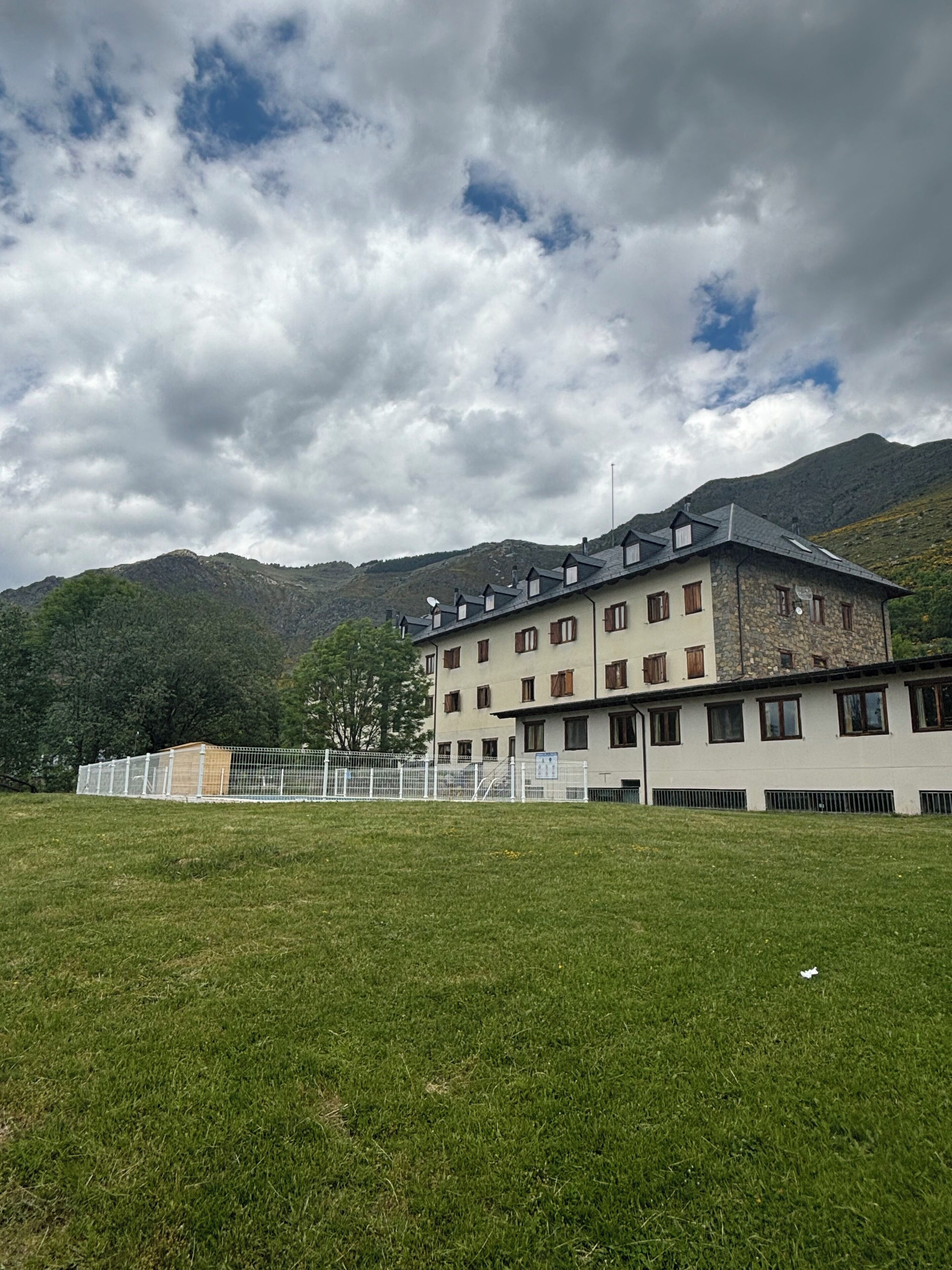 Un edificio con múltiples ventanas y techo inclinado, rodeado por un campo cubierto de césped y montañas al fondo.