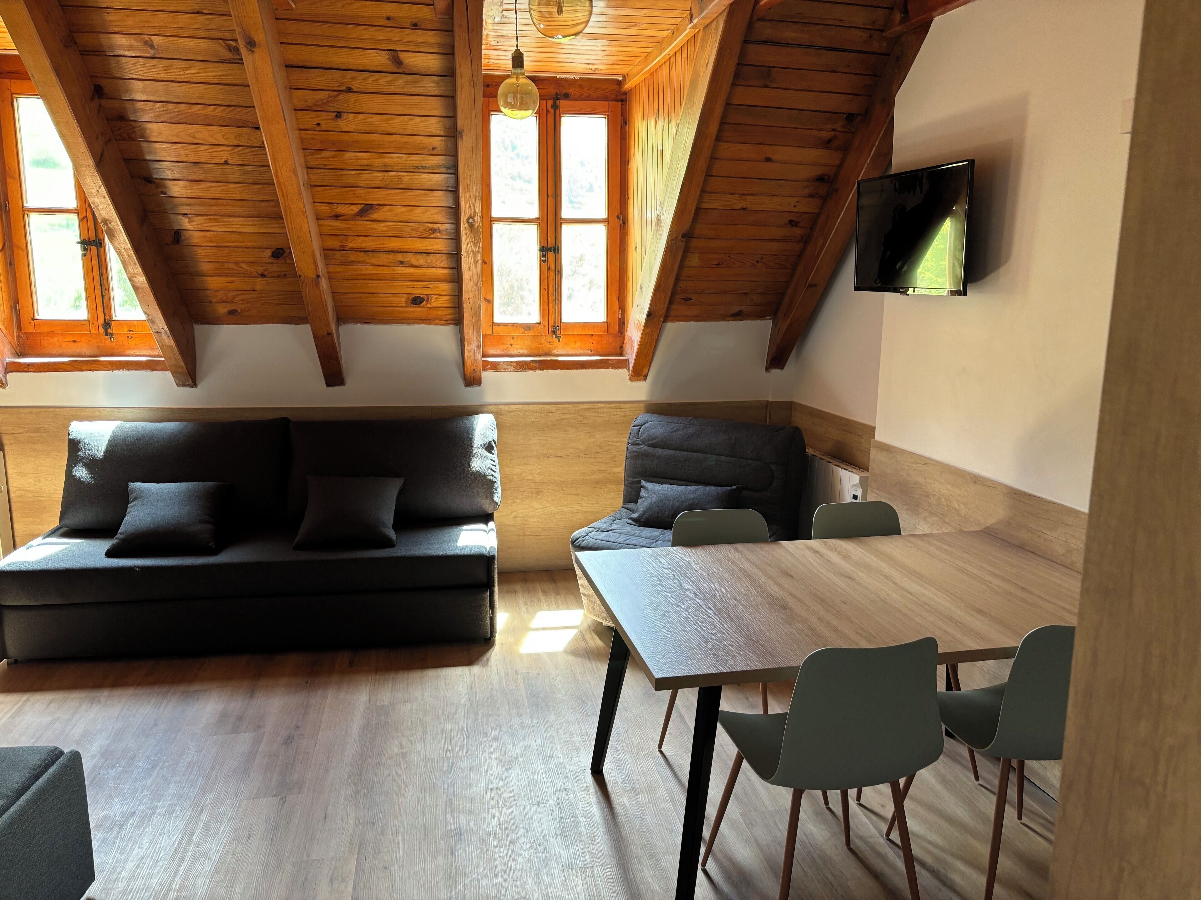Een gezellige loftkamer met houten plafond, een bank, een eettafel met stoelen, en een aan de muur bevestigde televisie.