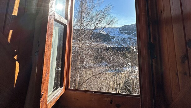 Una cabaña de madera con ventana que muestra un paisaje nevado y árboles desnudos.