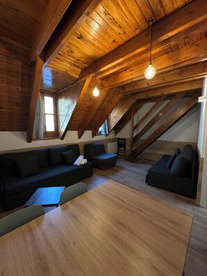 Habitación estilo loft con techo de madera, un sofá modular negro, una mesa de comedor de madera y una escalera que sube a un nivel superior.