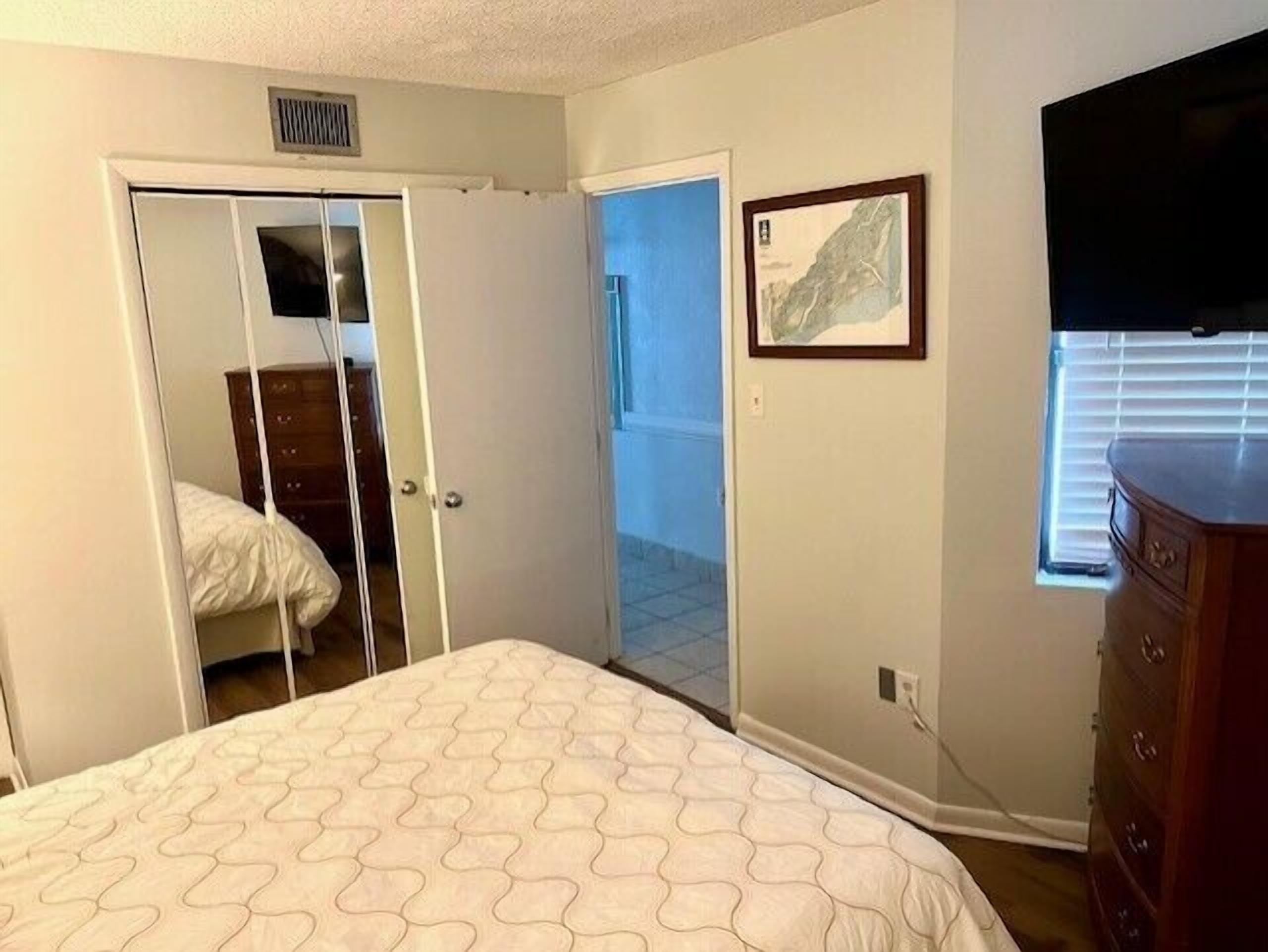 Une chambre à coucher comprenant un lit, une commode, un téléviseur, une porte donnant sur un balcon et un tableau encadré accroché au mur.