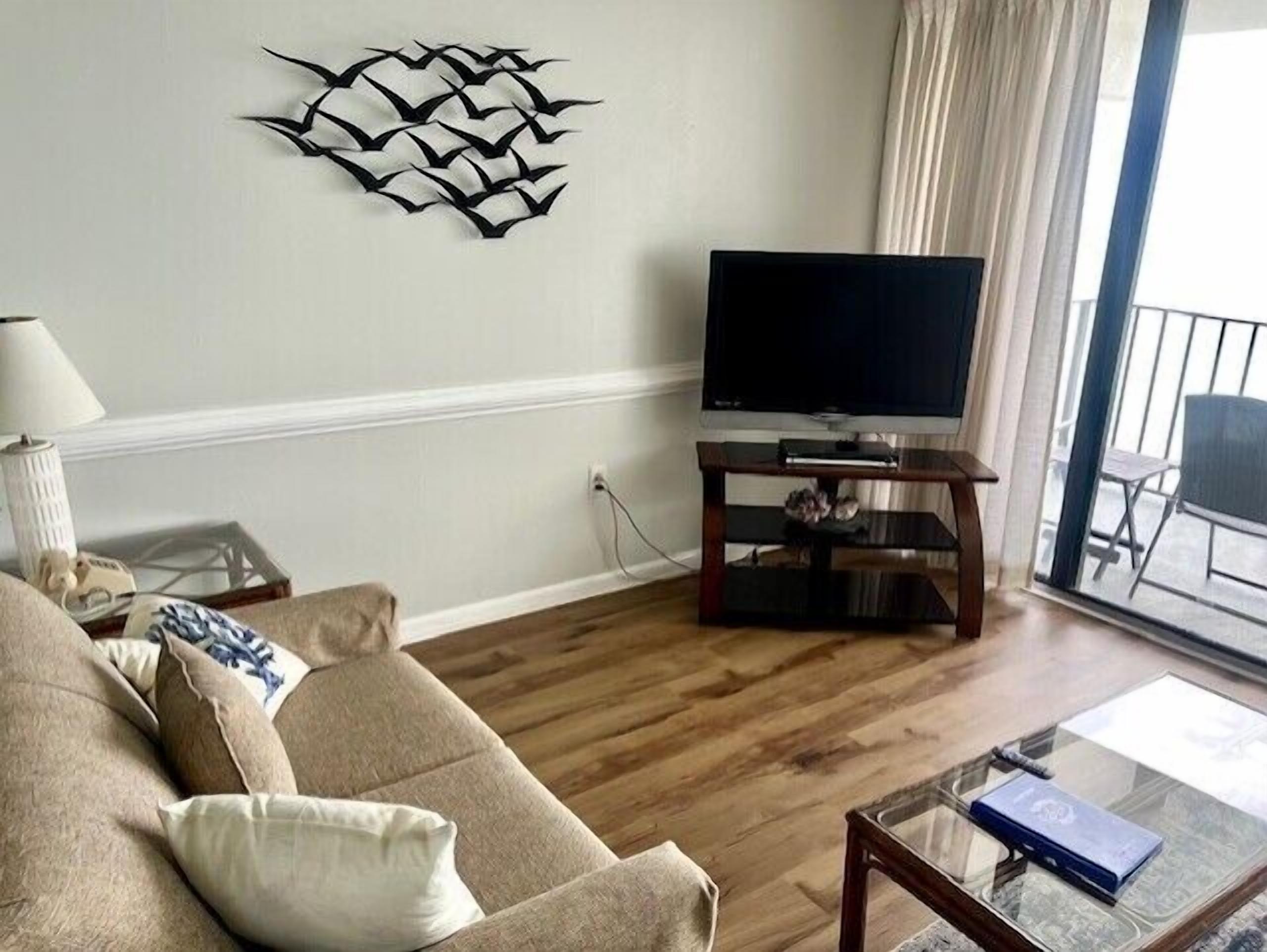A living room with a sofa, a glass coffee table, a television on a wooden stand, and a decorative wall piece.