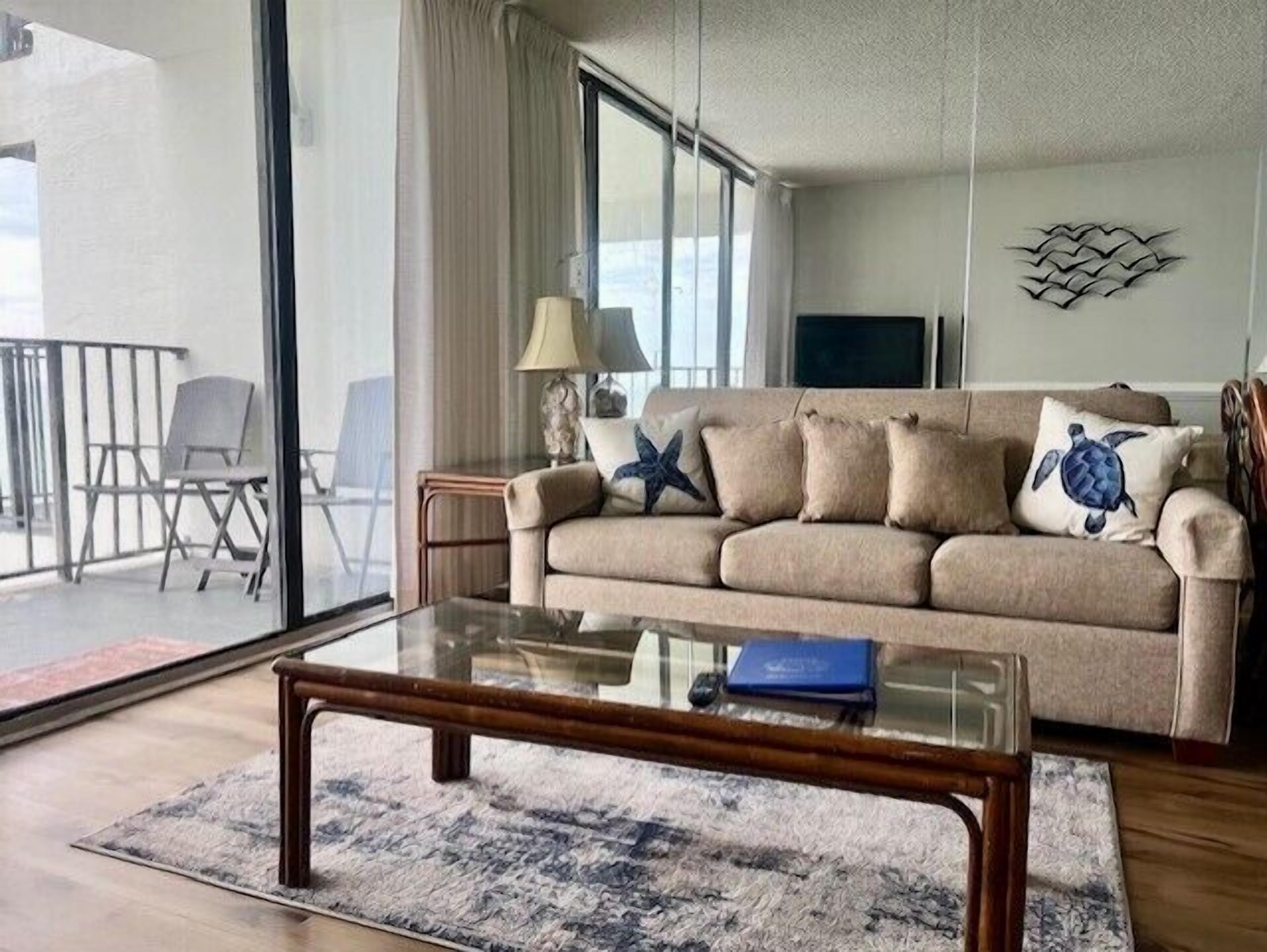 A living room with a beige sofa, a glass coffee table, a starfish pillow, and a turtle pillow. There is a TV on the wall and a balcony with a chair.