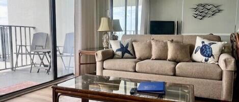 A living room with a beige sofa, a glass coffee table, a starfish pillow, and a turtle pillow. There is a TV on the wall and a balcony with a chair.