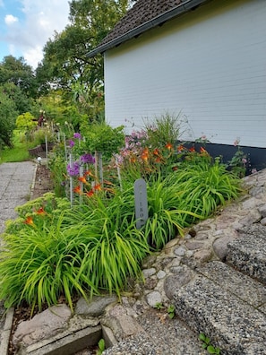 Un jardin où l’on trouve une grande variété de plantes et de fleurs, un sentier en pierre et un bâtiment blanc doté d’un toit foncé.