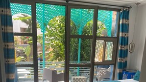 A room with a bed, two ottomans, a chair, and a view through large windows to an outdoor area with a green structure. - Private_vacation_home (El Jadida)
