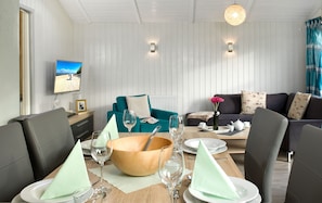 Un coin repas avec une table en bois dressée pour quatre personnes, entourée de fauteuils en cuir foncé. On y trouve un canapé turquoise et un canapé gris foncé agrémenté de coussins à motifs. Un téléviseur diffusant une scène de plage est fixé au mur.