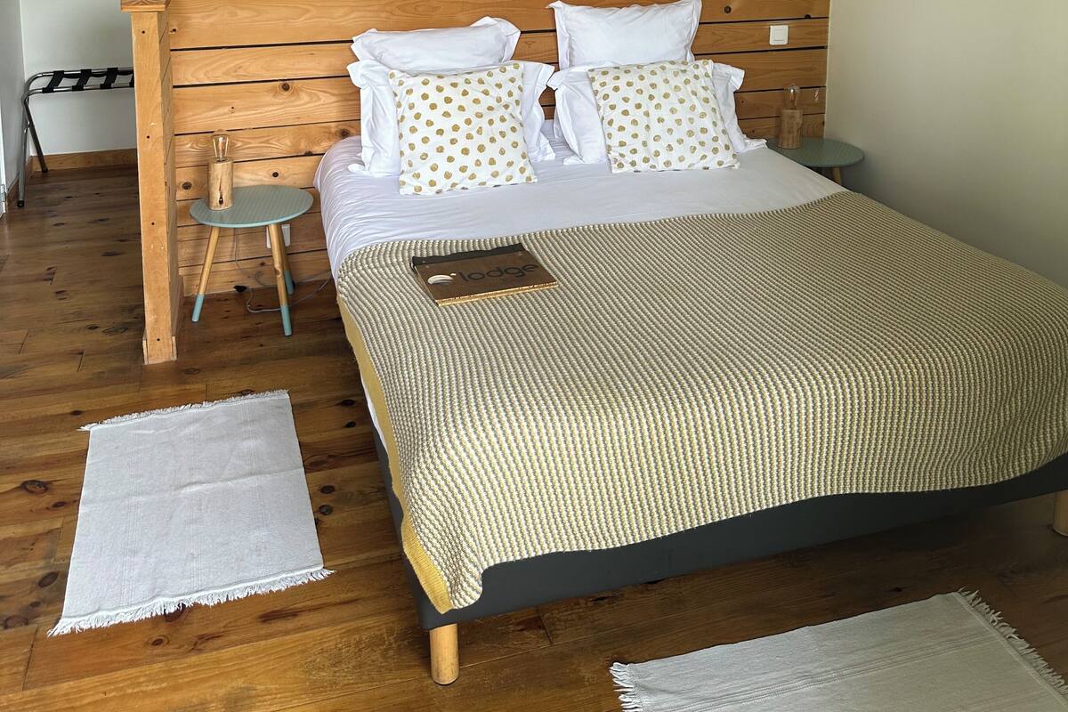 A bedroom with a wooden headboard, a bed with white and polka-dotted pillows, a bedside table, and a small stool.