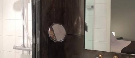 A modern bathroom with a glass shower enclosure, a round mirror, a sink with a faucet, and toiletries on the counter.