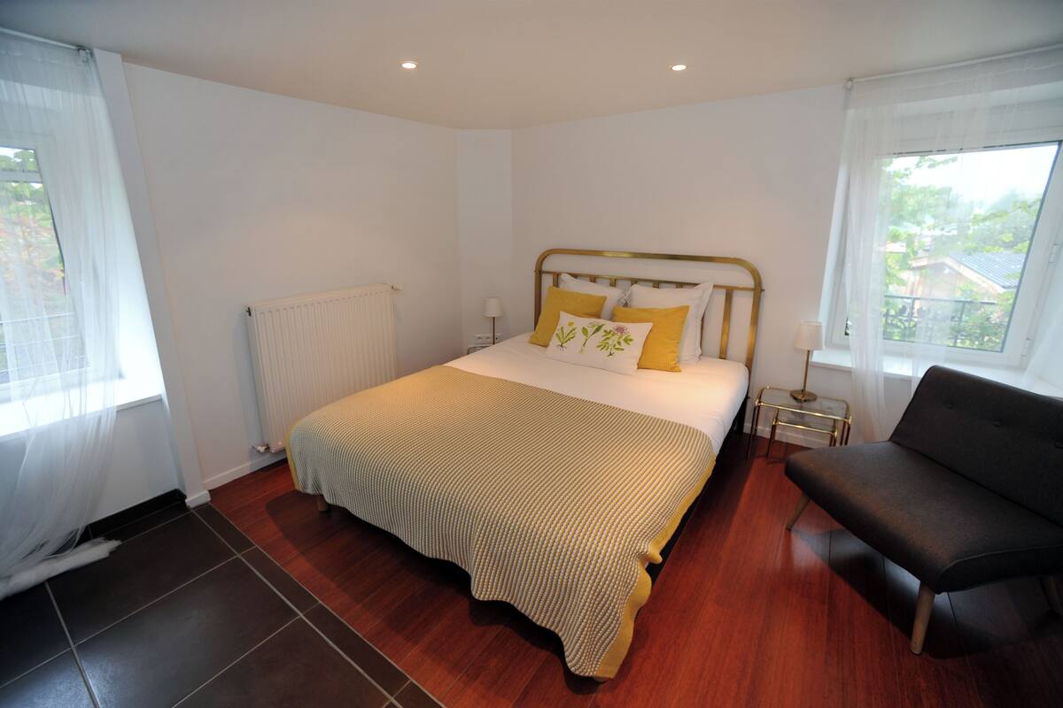 A bedroom with a bed, bedside tables, a radiator, a window with curtains, and a sofa.