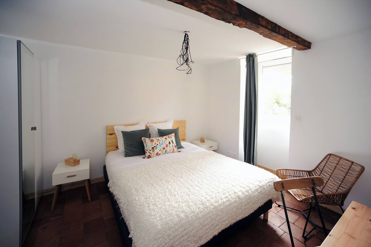 A bedroom with a large bed, a wooden nightstand, a wicker chair, and a window with curtains.