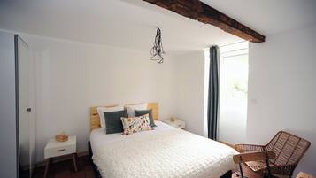 A bedroom with a large bed, a wooden nightstand, a wicker chair, and a window with curtains.