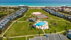 Un complexe hôtelier comprenant plusieurs bâtiments, une piscine centrale, des courts de tennis et offrant une vue sur la plage.