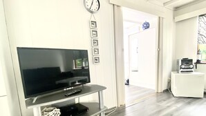 Una sala de estar moderna con un televisor de pantalla plana sobre un soporte metálico, un reloj en la pared y una puerta que da a otra habitación.