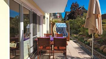 Um pátio com mesa e cadeiras, um guarda-sol listrado e uma porta de vidro que dá para um jardim.