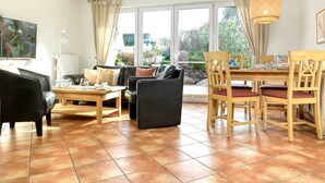 A modern living room with a sofa, a coffee table, and a dining table with chairs. Large windows provide a view of the outdoors. - App. Wellenreiter is looking forward to your visit! (Insel Poel / OT Schwarzer Busch)