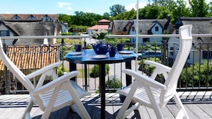 Un balcon avec une table dressée pour deux personnes, des chaises blanches, un parasol coloré et une vue sur des maisons aux toits de chaume.