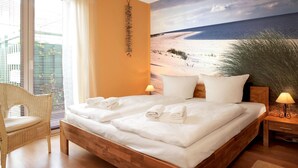 A bedroom with a wooden bed, white linens, a wicker chair, a nightstand with a lamp, and a beach mural on the wall. - App. Leuchtfeuer - Vacation right on the beach! (Insel Poel / OT Schwarzer Busch)
