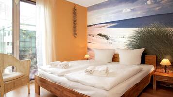 A bedroom with a wooden bed, white linens, a wicker chair, a nightstand with a lamp, and a beach mural on the wall.