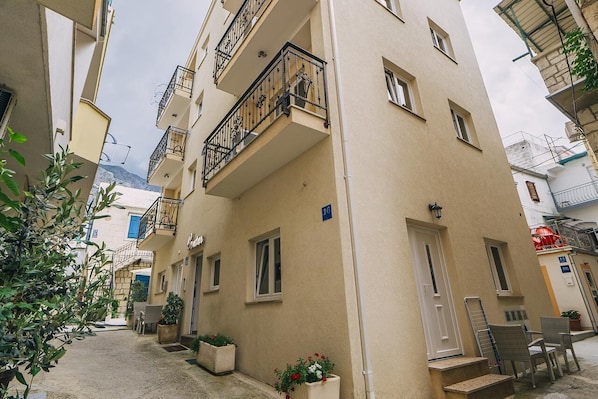 A narrow street with buildings on either side, featuring balconies with metal railings, a few potted plants, and a visible door with steps leading to an entrance.
