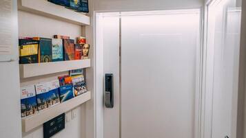 Un pasillo de hotel con una puerta blanca, una cerradura digital y estantes con libros y revistas.