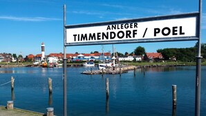 Eine hölzerne Anlegestelle mit einem Schild, auf dem "Anleger Timmendorf / Poel" steht, in einer ruhigen Wasserlandschaft mit Häusern und einem Leuchtturm im Hintergrund.