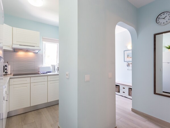 A modern kitchen with white cabinets, a built-in oven, and a window. There is a hallway leading to a dining area with a clock on the wall.