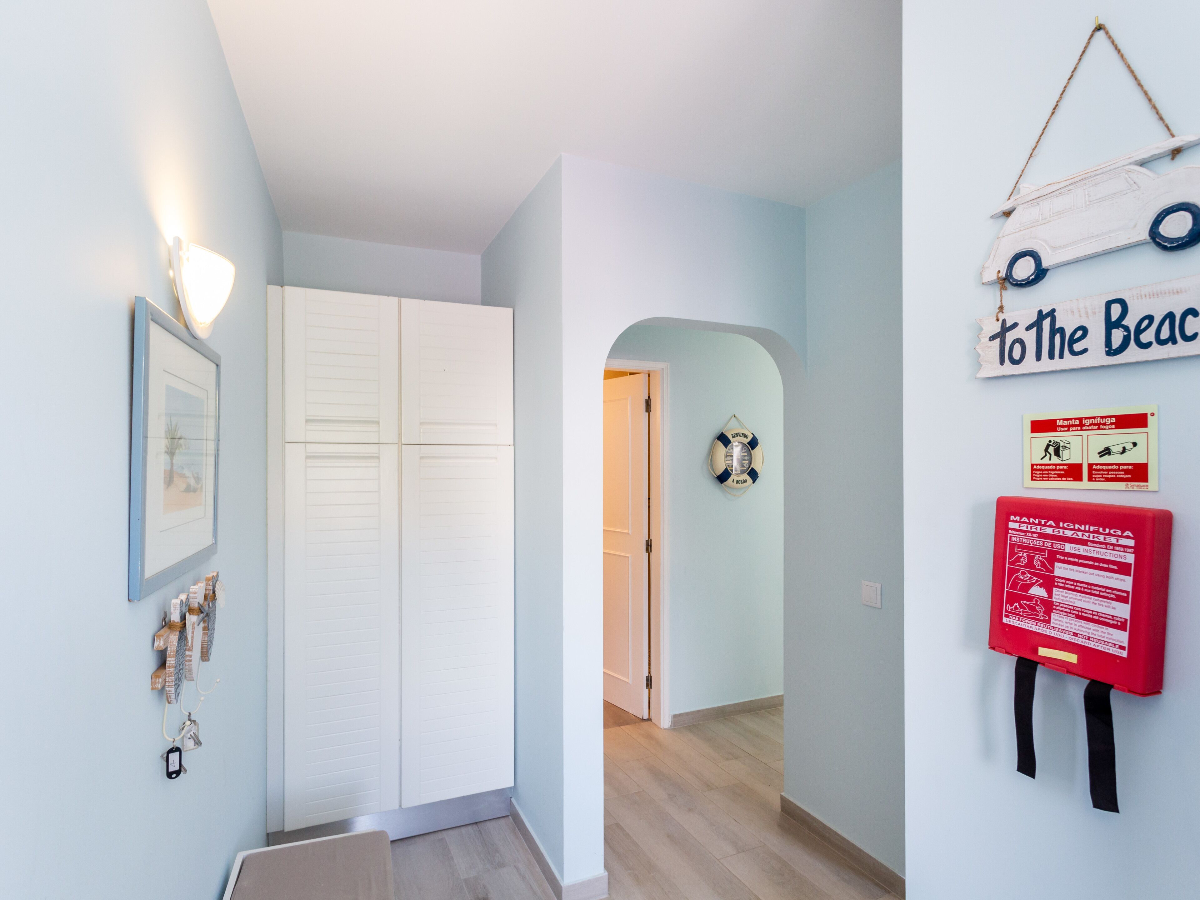 Un couloir avec une armoire blanche, un tableau encadré, un gilet de sauvetage et un panneau indiquant « Vers la plage ».
