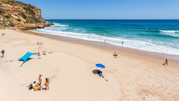 A sandy beach with a rocky cliff, clear blue water, and people sunbathing and swimming. - Casa Pedro - Burgau - Unique Converted Fisherman's Cottage (Budens)