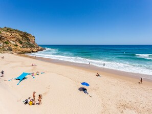 A sandy beach with a rocky cliff, clear blue water, and people sunbathing and swimming.