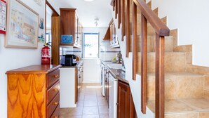 A narrow kitchen with wooden cabinets, a coffee maker, and a fire extinguisher. There is a staircase with wooden railings leading to another level. - Casa Pedro - Burgau - Unique Converted Fisherman's Cottage (Budens)