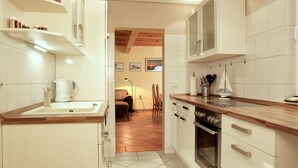 A narrow kitchen with white cabinets, a wooden countertop, and a tiled floor. There is a coffee maker on the counter and a chair in the background.