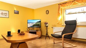 A living room with a wooden table, a TV on a stand, a chair, and a window with curtains. - Apt. Dwarslöper - Peaceful holiday apartment in Kirchdorf in the owner's house. (Insel Poel / OT Kirchdorf)