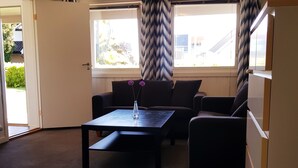 A room with a sofa, a coffee table, and a refrigerator. There are windows with curtains, and a door leading outside. - StayPlus Central Apartment With Garden (Kristiansand)