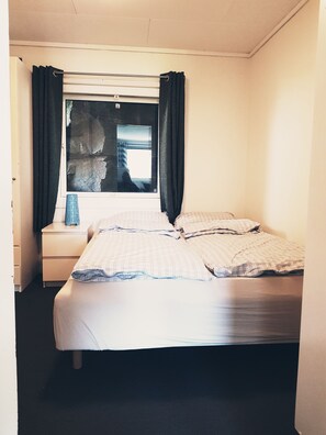 A single bed with a checkered duvet, a bedside table with a lamp, and a window with curtains.